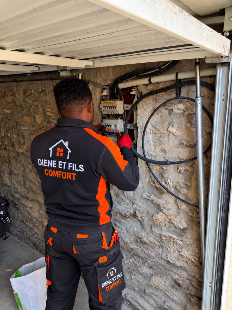 Électricien Diene et Fils Comfort raccordant un tableau électrique dans un garage, câbles et gaines rangés le long d’un mur en pierre